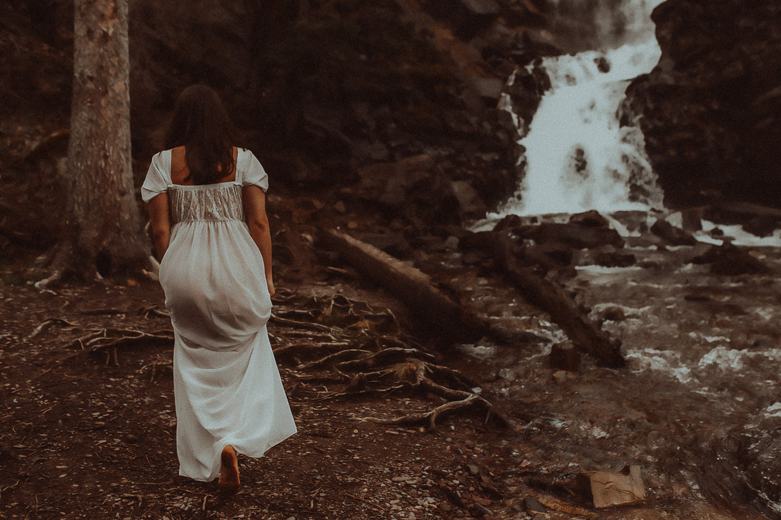A woman in a white gown walks along a river with a waterfall