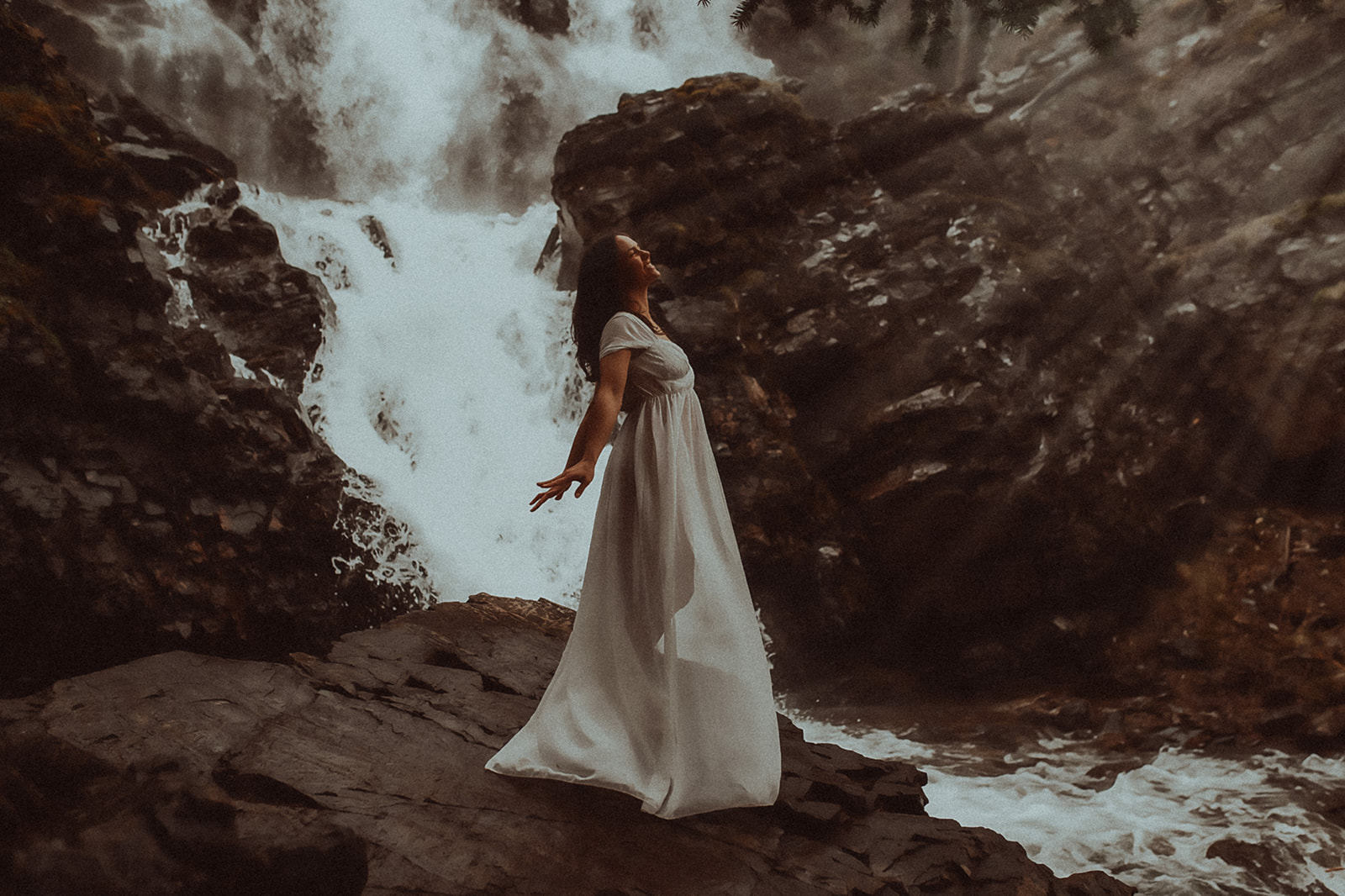 A happy woman in a white gown throws her hands back under a waterfall after visiting hair salons in Montana