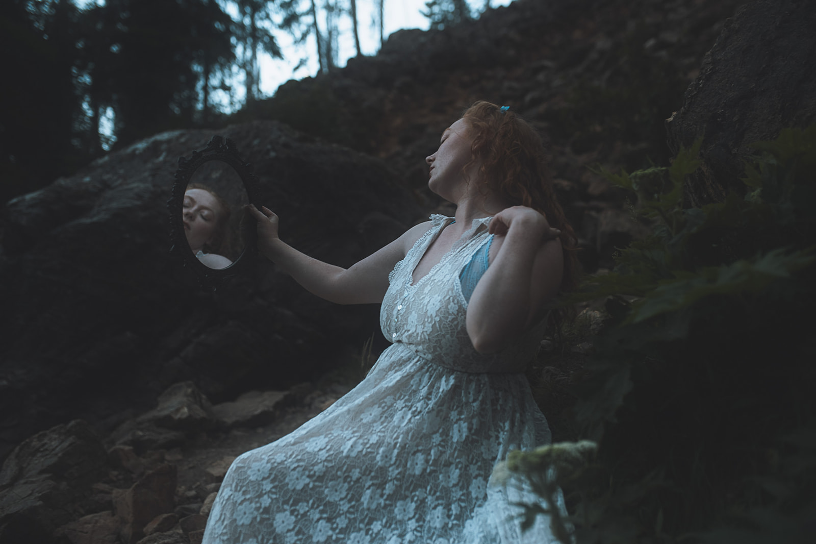 A woman in a lace dress sits on a rock wall admiring herself in a handheld mirror