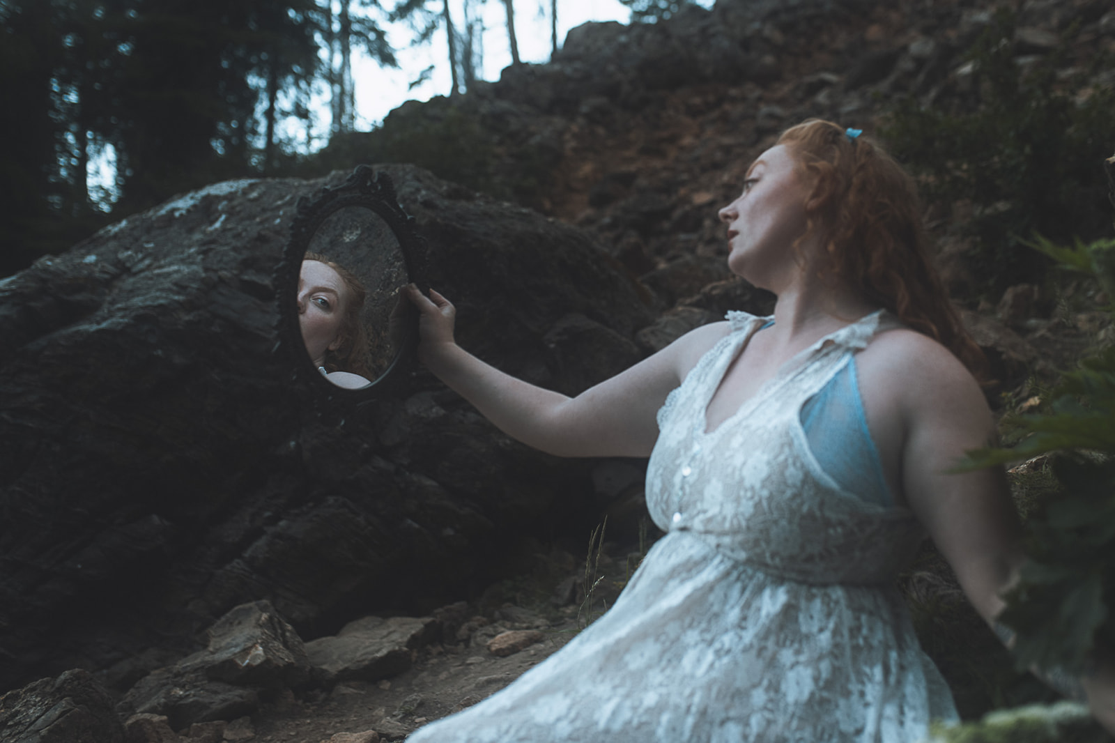 A woman with red hair admires herself in a hand held mirror in a forest