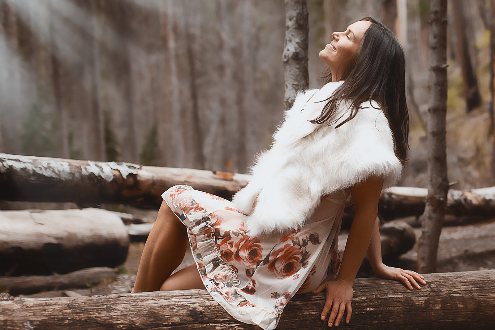 A woman smiles while wearing a fur shawl and pink slip and sitting on a log in a forest