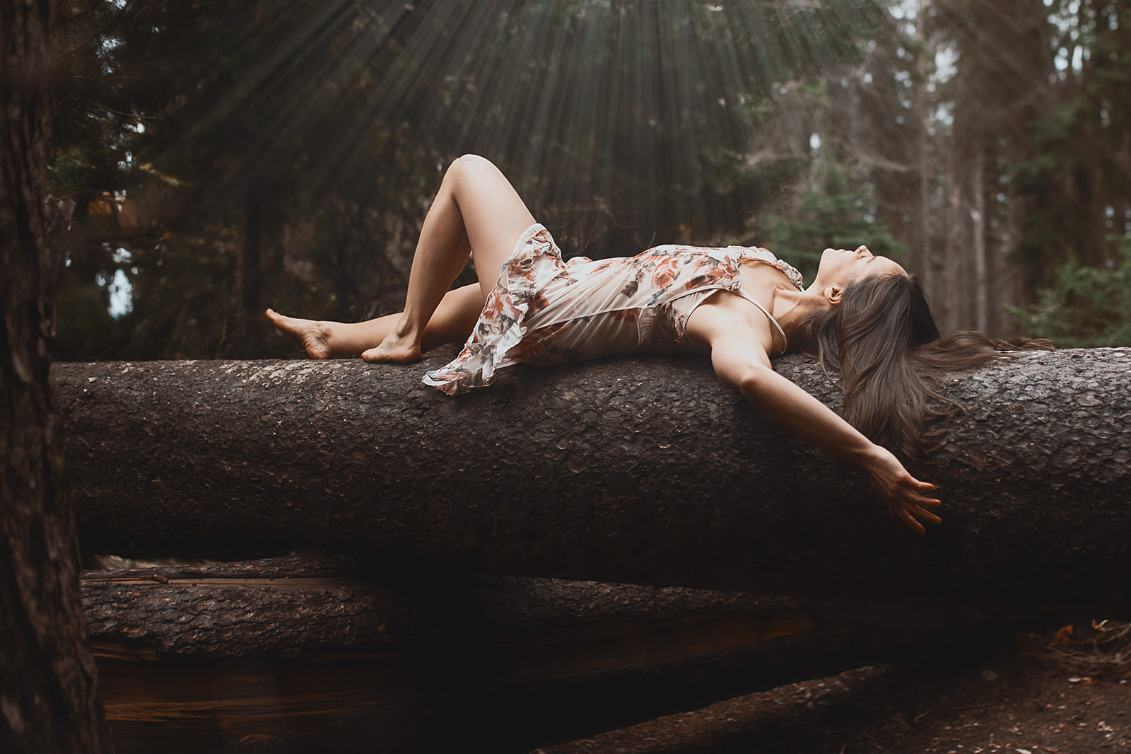 A woman in a pink slip basks in the sun beams in a forest across a fallen tree after some Missoula Pilates