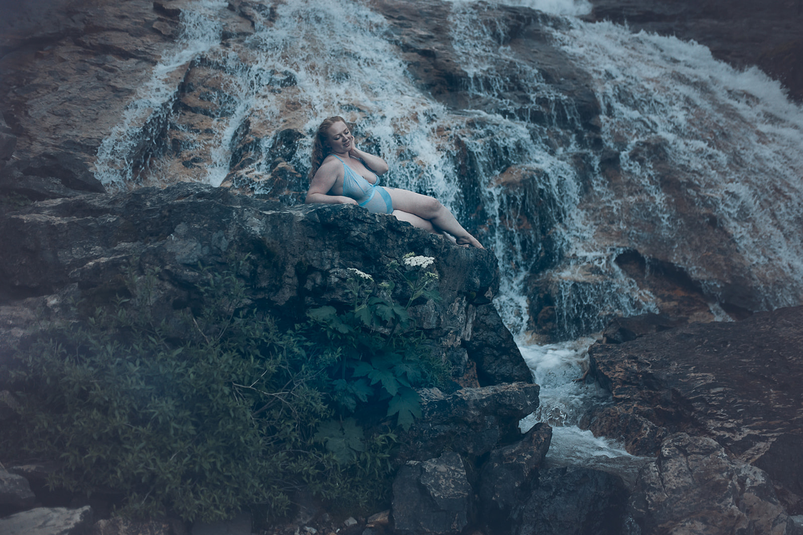 A woman lounges on a boulder by a waterfall in blue lingerie