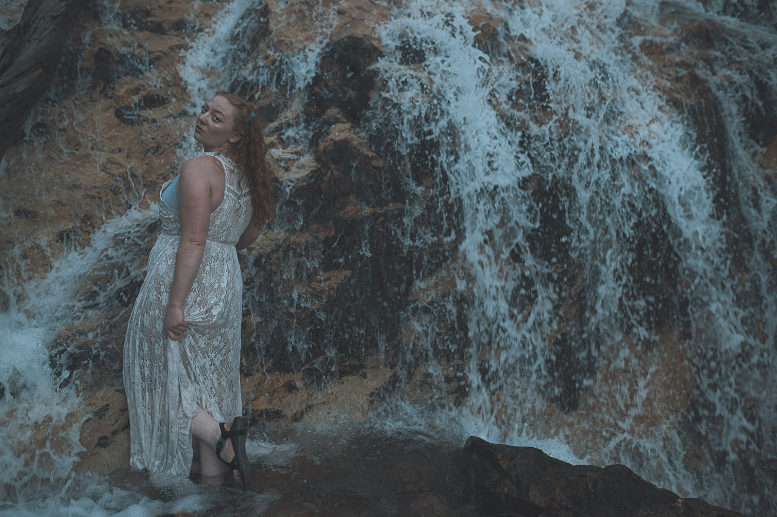 A woman with red hair walks in the water below a waterfall in a lace dress after visiting hair salons in Kalispell, MT