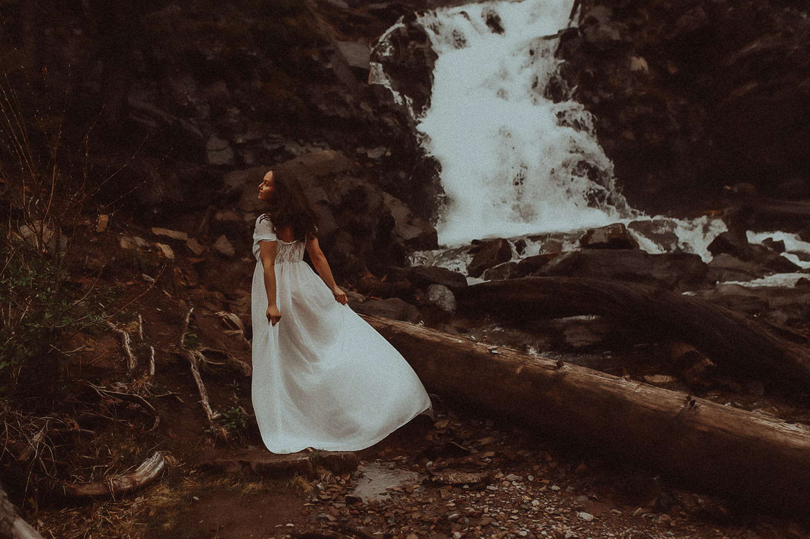 A woman in a white gown walks through a trail by a waterfall after some Helena Yoga