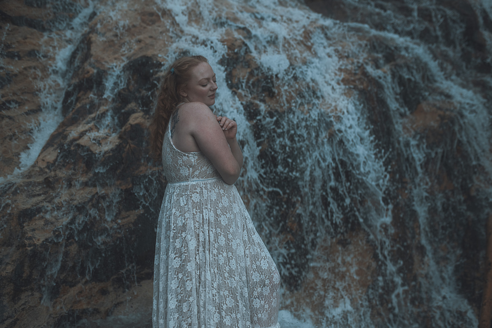 A woman hugs herself while exploring a waterfall in a white lace dress