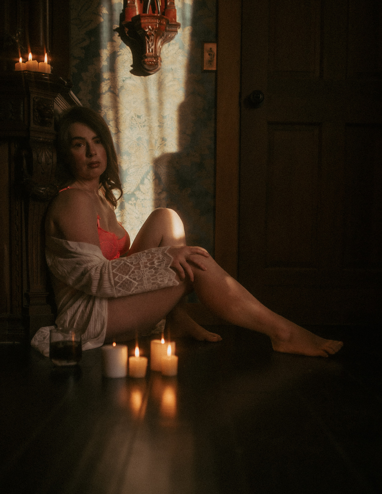 A woman in hot pink lingerie and a white coverup sits against a dresser with candles