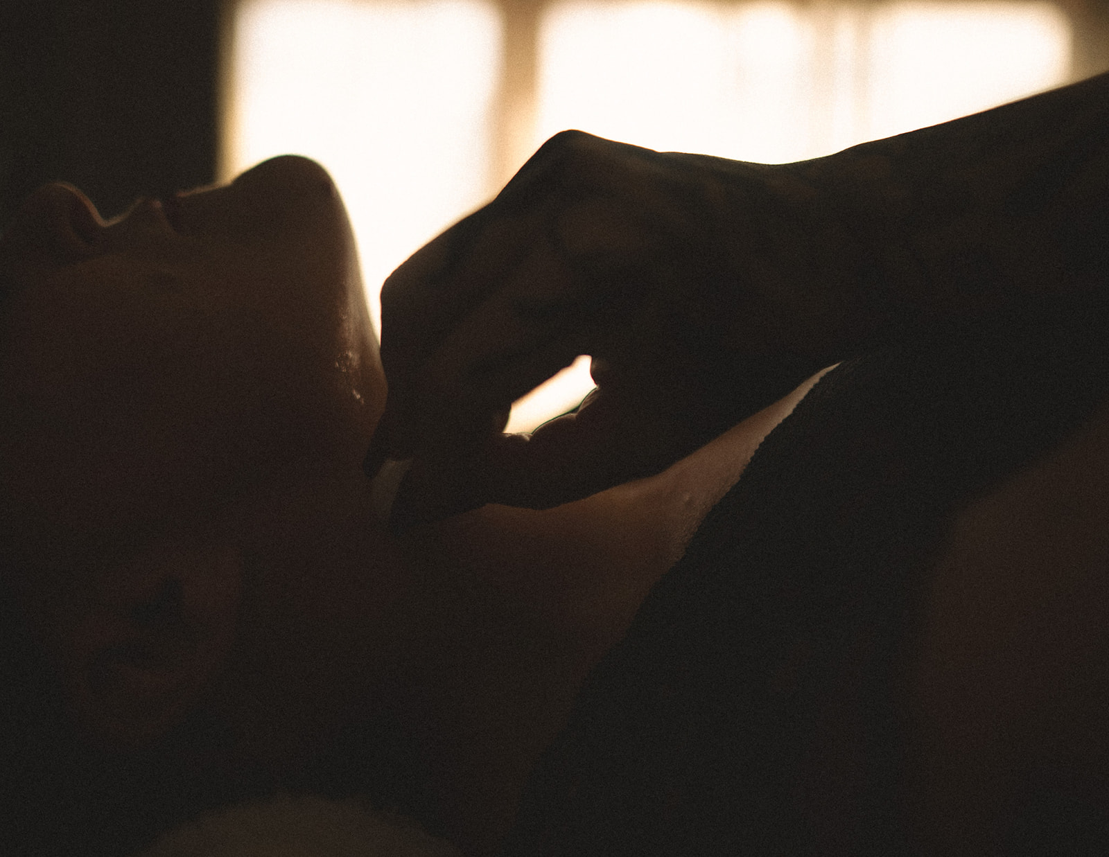 A woman rubs ice cubes on her neck while laying on a bed in black lingerie