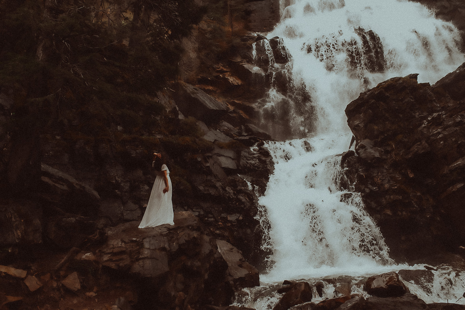A woman in a long white dress admires a toll waterfall while climbing up it after some yoga in Hamilton, MT