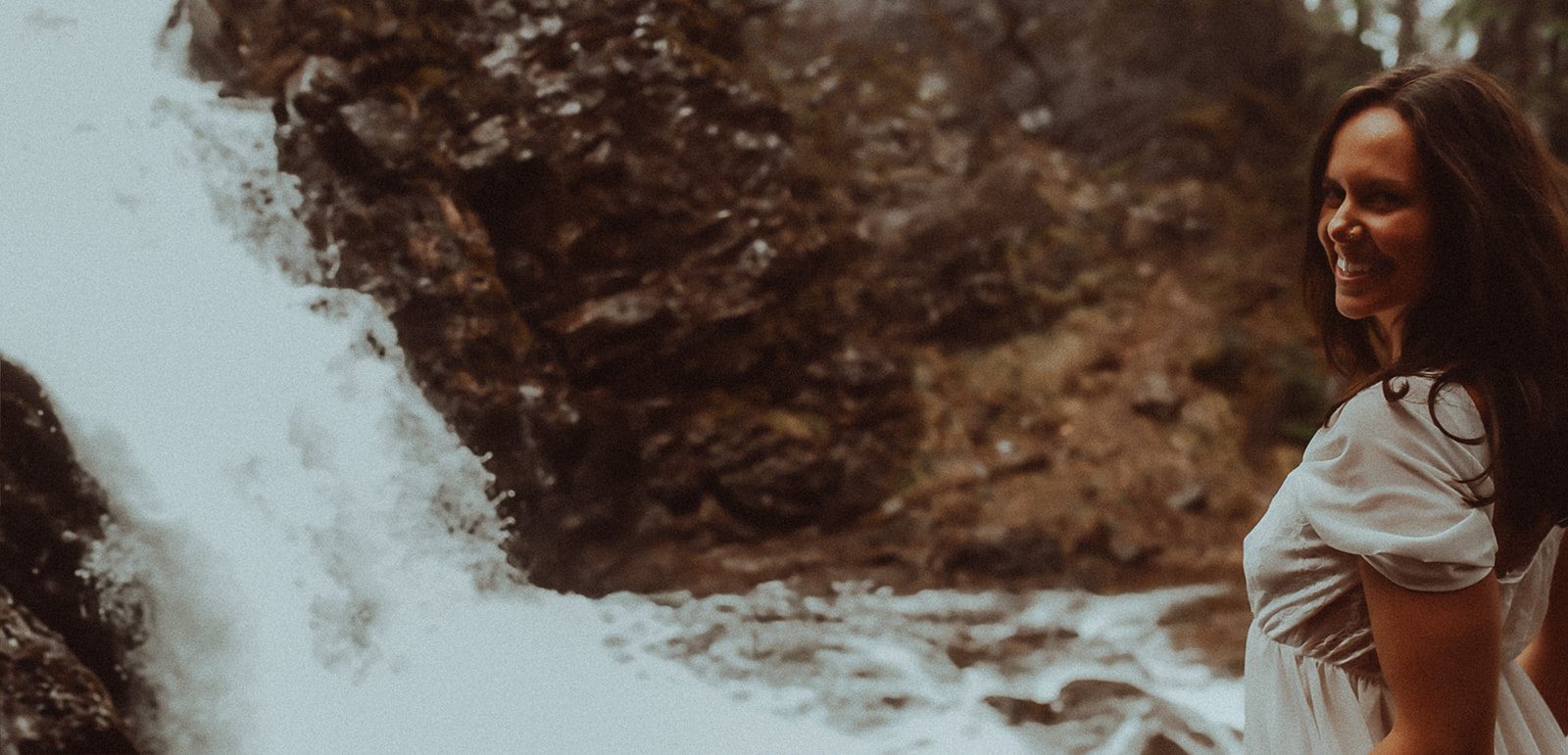 A woman smiles over her shoulder in a white dress at the base of a waterfall after some yoga in Hamilton, MT