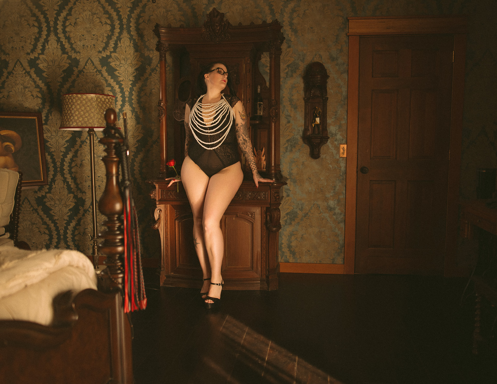 A woman leans against a vintage hutch in black lingerie, pearls and holding a red rose after exploring lash extensions in Missoula