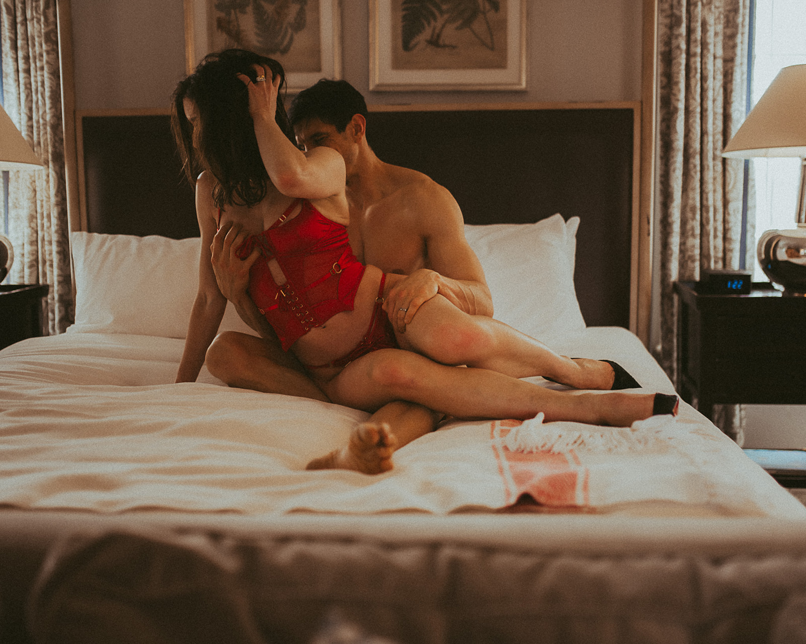 A couple snuggle on a bed while the woman wears a red corset