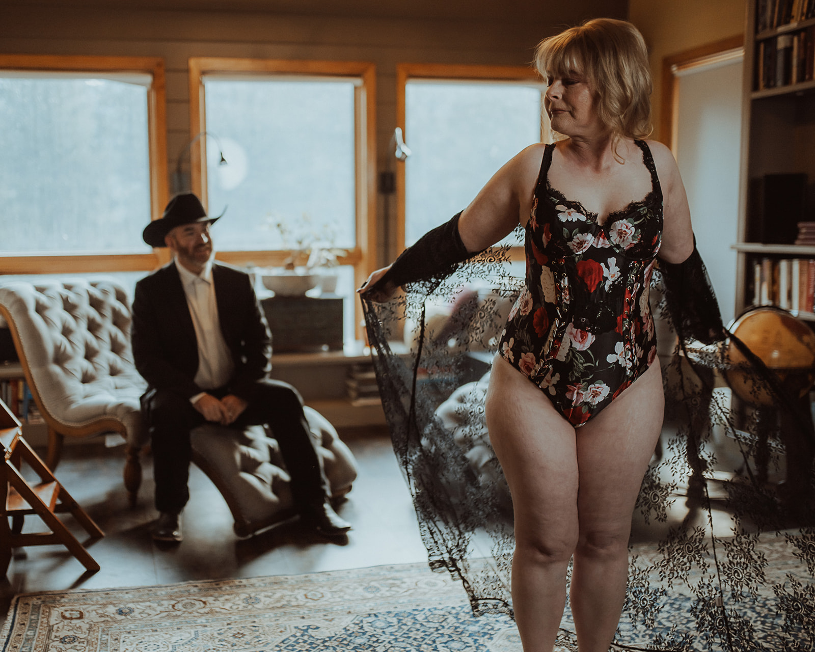 A woman in a flower print corset and lace train shows off to her husband sitting on a chair across the room