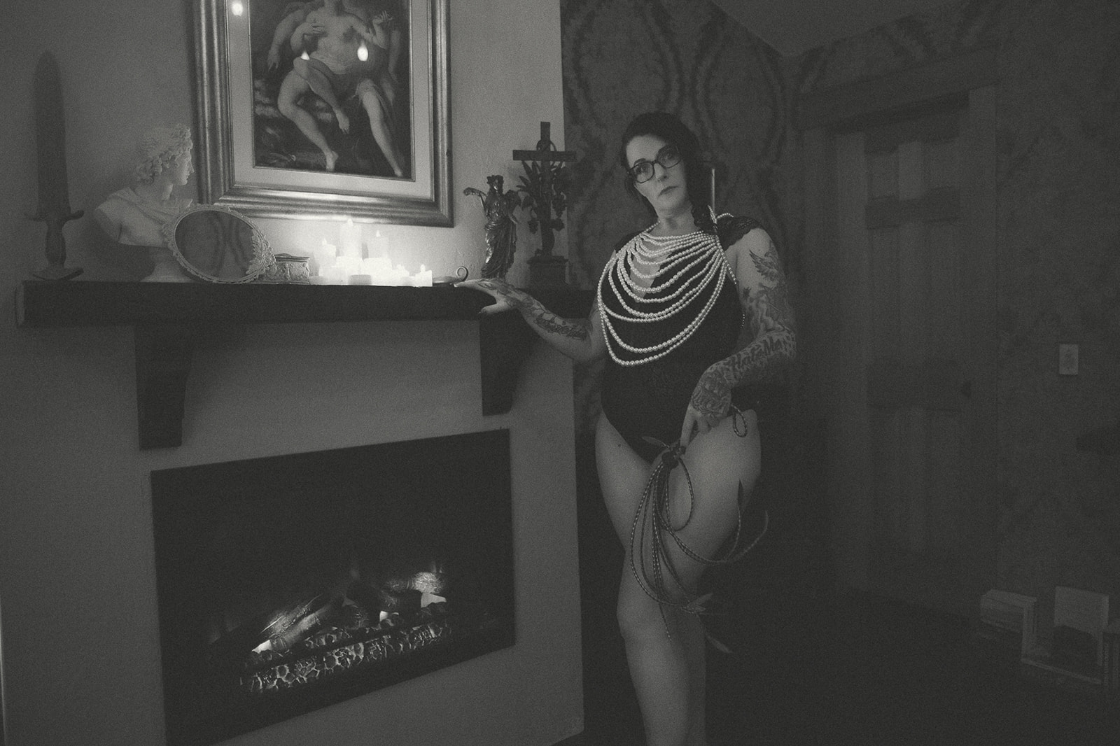 A woman covered in pearls holds a short whip while standing by a fireplace in a lace bodysuit after finding lash extensions in Kalispell